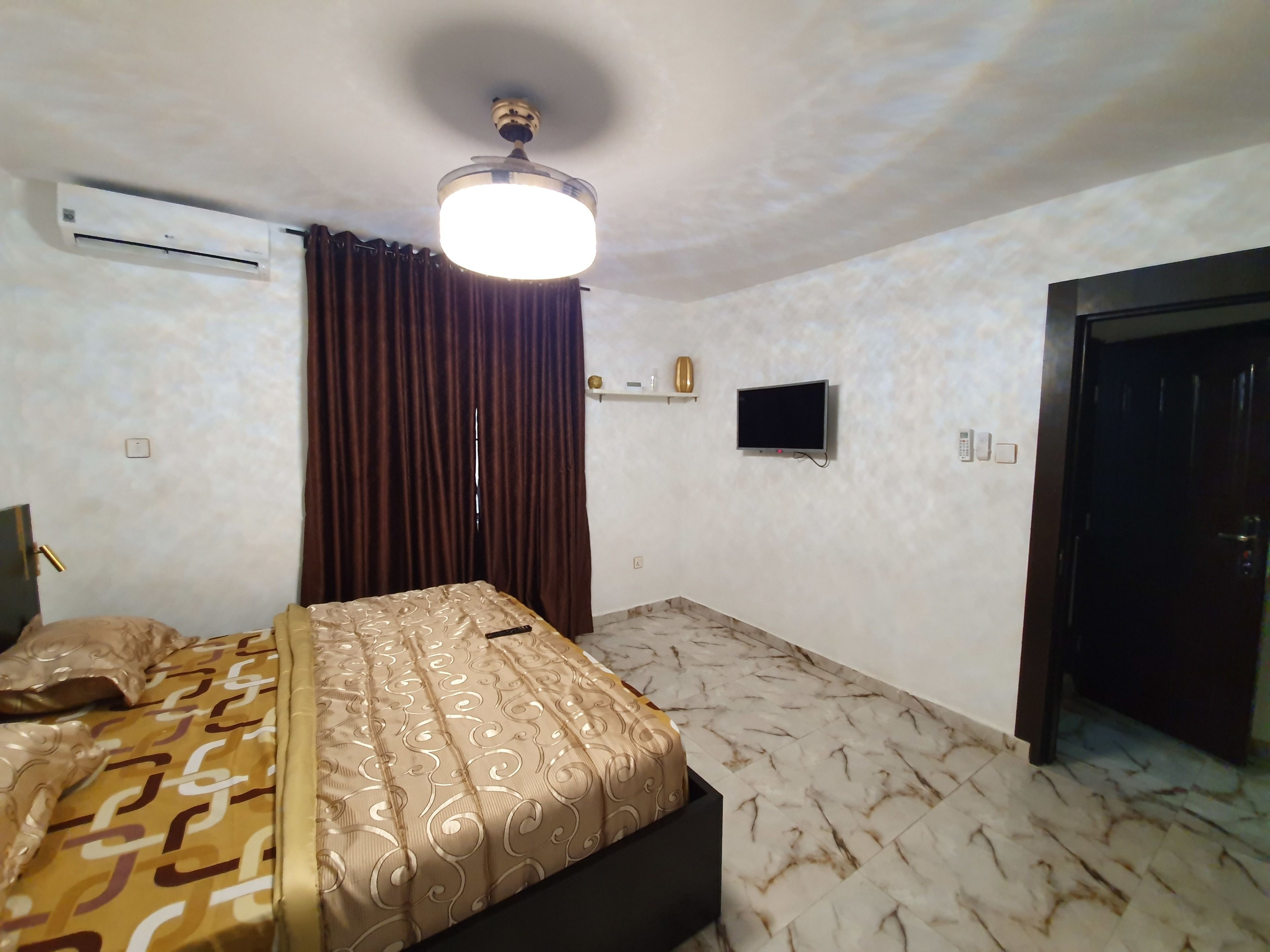 A hotel room with a bed, a ceiling fan, an air conditioning unit, a television, a door, and a marble-patterned floor.