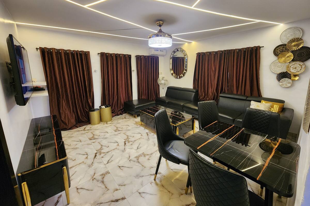 Una sala de estar moderna con un sofá negro, una mesa de comedor con sillas, piso de mármol y una lámpara de techo.
