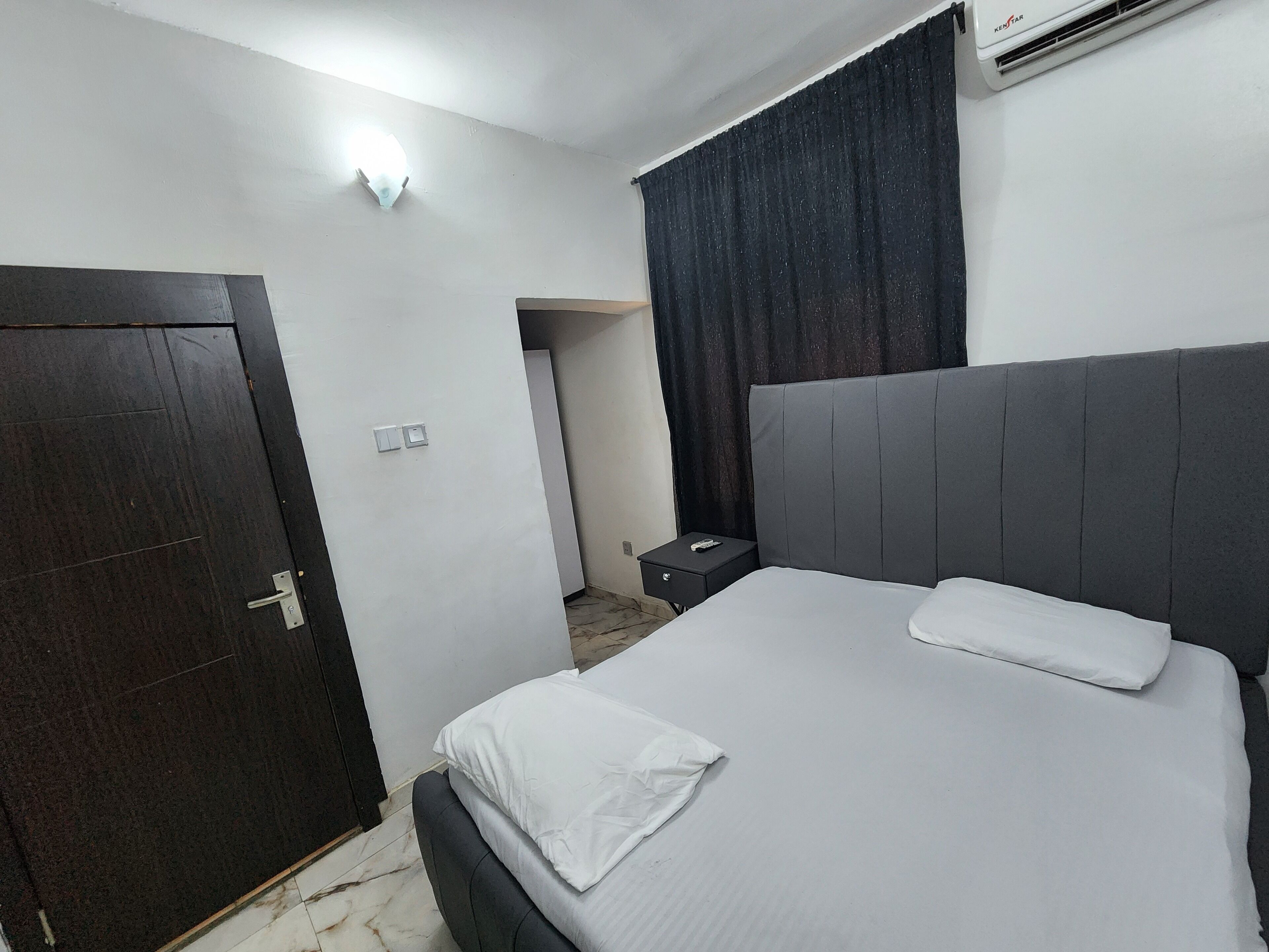 A hotel room with a bed, a nightstand, a dark wooden door, a light switch, and an air conditioning unit.