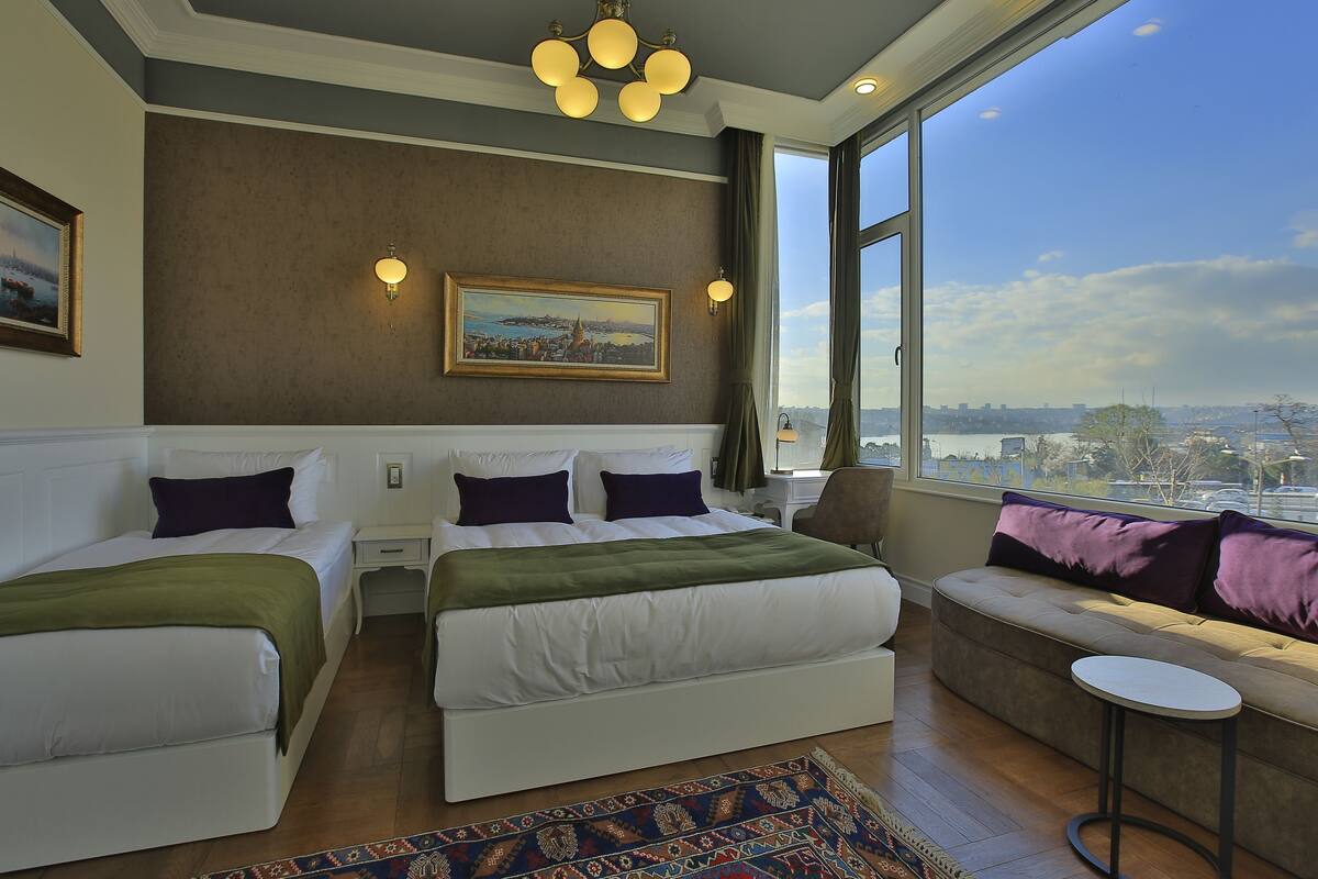 A hotel room with two beds, a sofa, a small table, and a large window offering a city view.