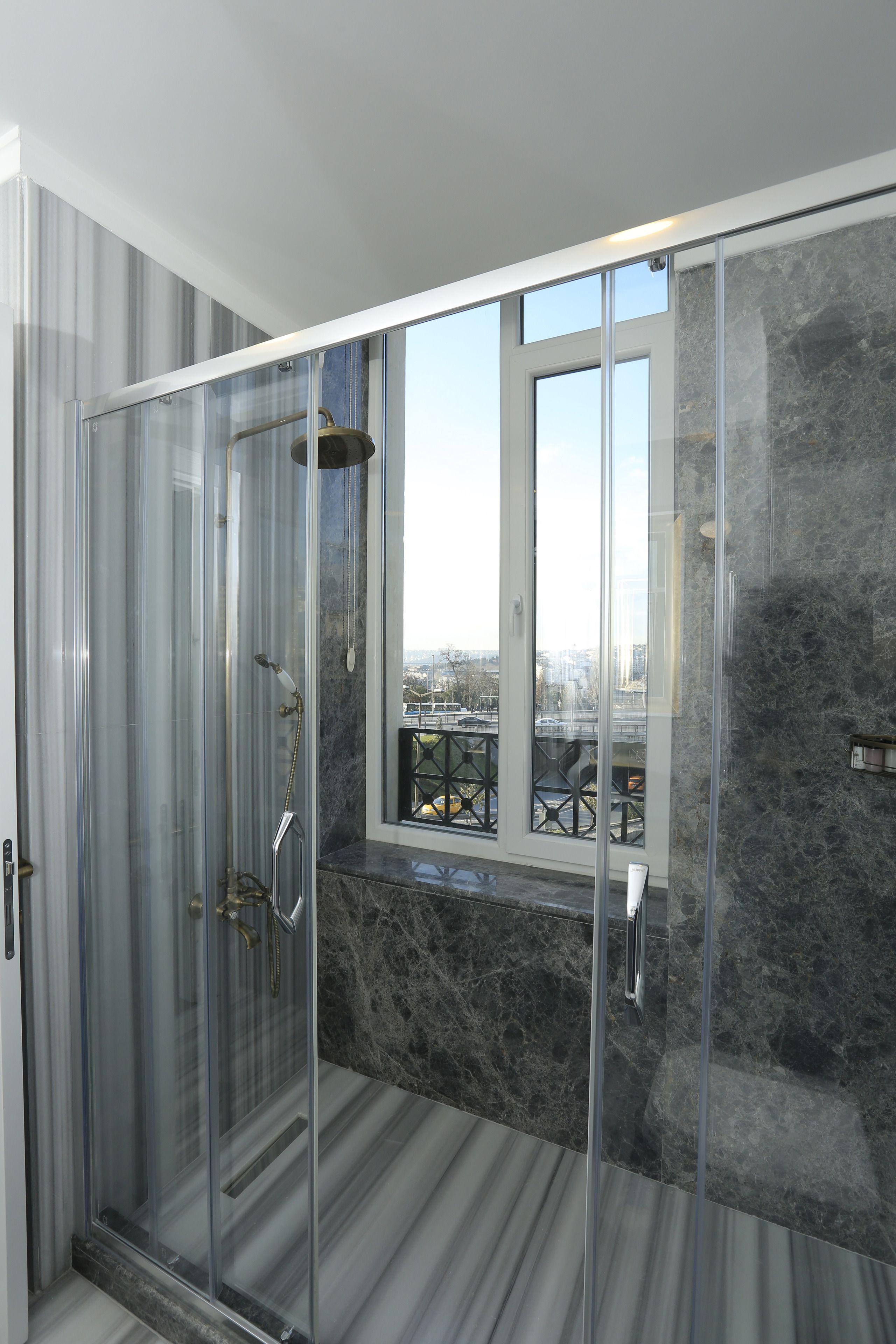 A modern shower with glass doors, a marble floor, and a view of the city through a window.
