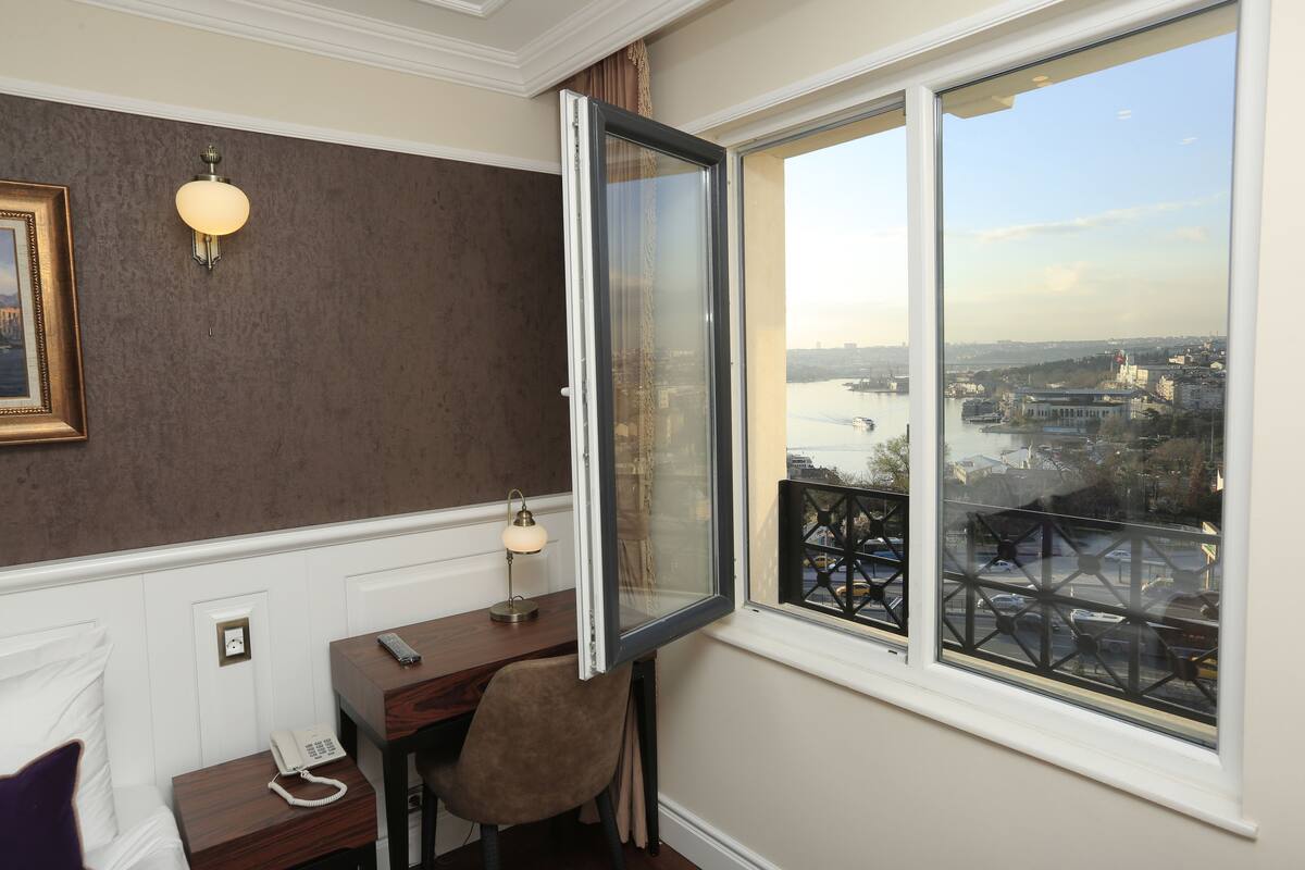 A room with a desk, chair, and a telephone. A window with a view of a cityscape and a bridge is visible.