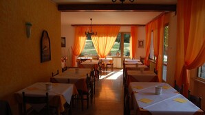 Un restaurante con varias mesas y sillas, un candelabro y ventanas amplias con cortinas naranjas.