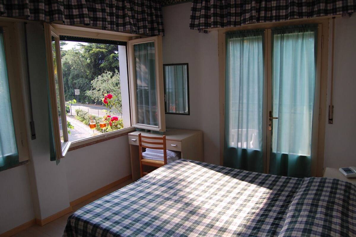 Een slaapkamer met een bed, een bureau en een stoel. Er is een raam met gordijnen en uitzicht op een tuin.