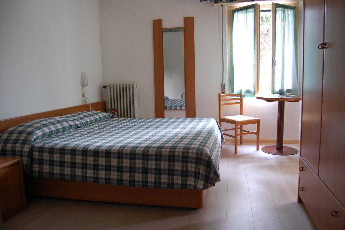 Een slaapkamer met een bed, een stoel, een spiegel, een radiator en een raam met gordijnen.