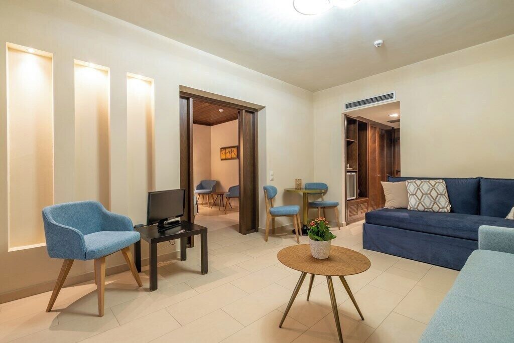 A modern living room with a sofa, two armchairs, a coffee table, and a TV. There are doors leading to other rooms.