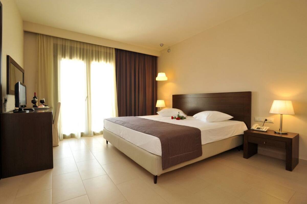 Ein Hotelzimmer mit einem großen Bett, zwei Nachttischlampen, einem Nachttisch, einem Schreibtisch mit Telefon und einem Fenster mit Vorhängen.
