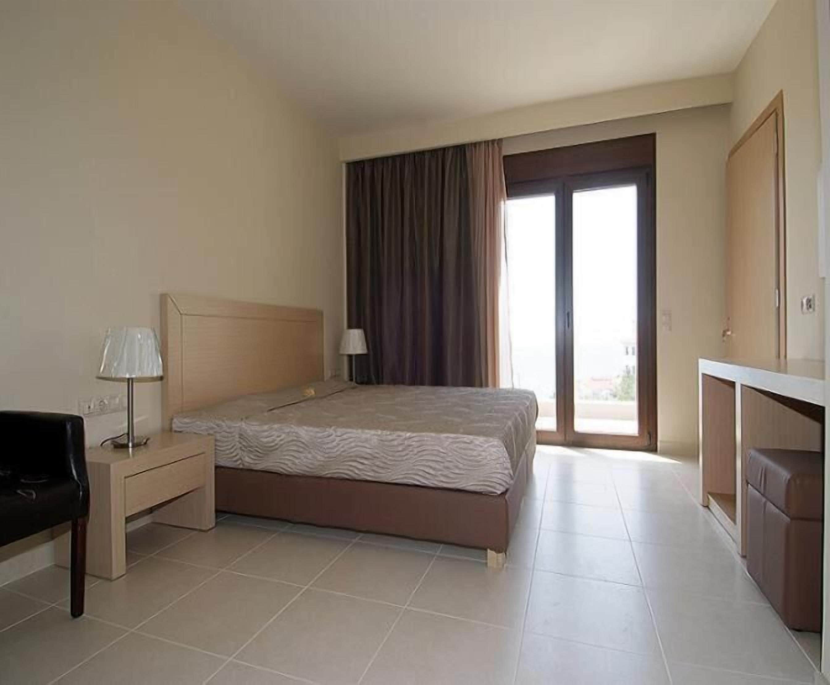 Habitación de hotel con cama, escritorio con lámpara, silla, armario y ventana grande con cortinas.
