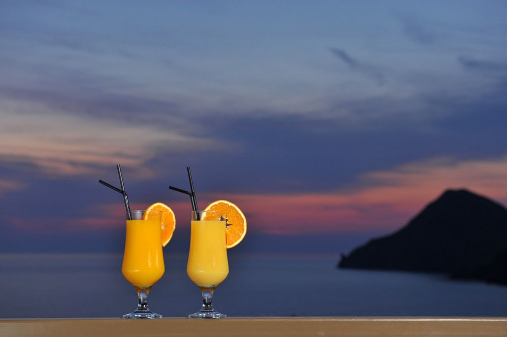 Two glasses of orange juice with straws and orange slices on a balcony with a view of a sunset and a distant island.