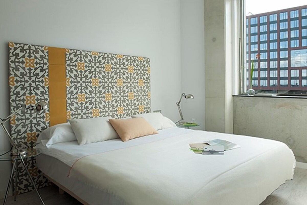 Una cama con cabecera estampada, una lámpara de noche y una ventana con vistas a un edificio.