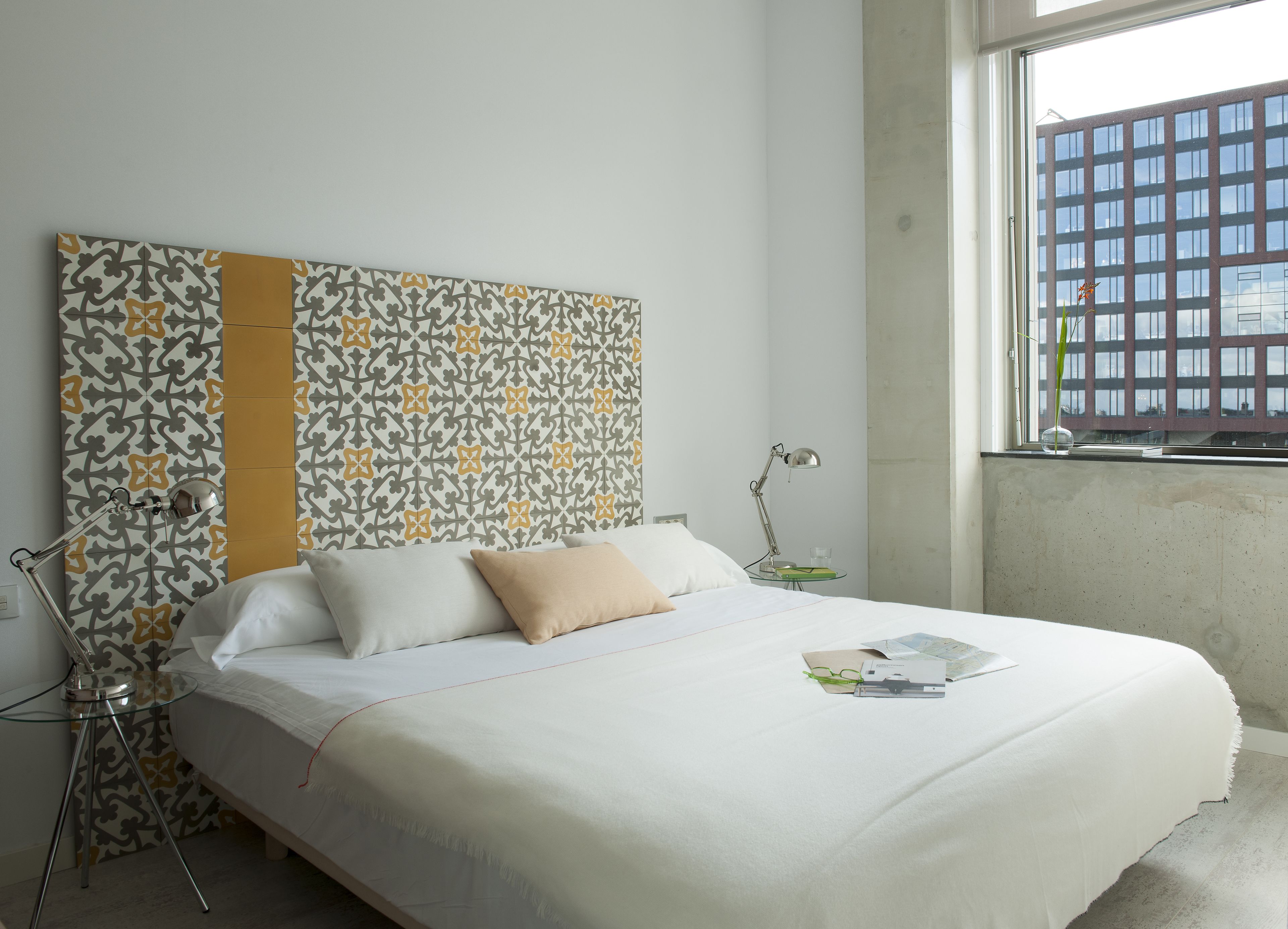 Cama bem arrumada com roupa de cama branca e travesseiro bege, mesa de cabeceira com abajur, e parede estampada.