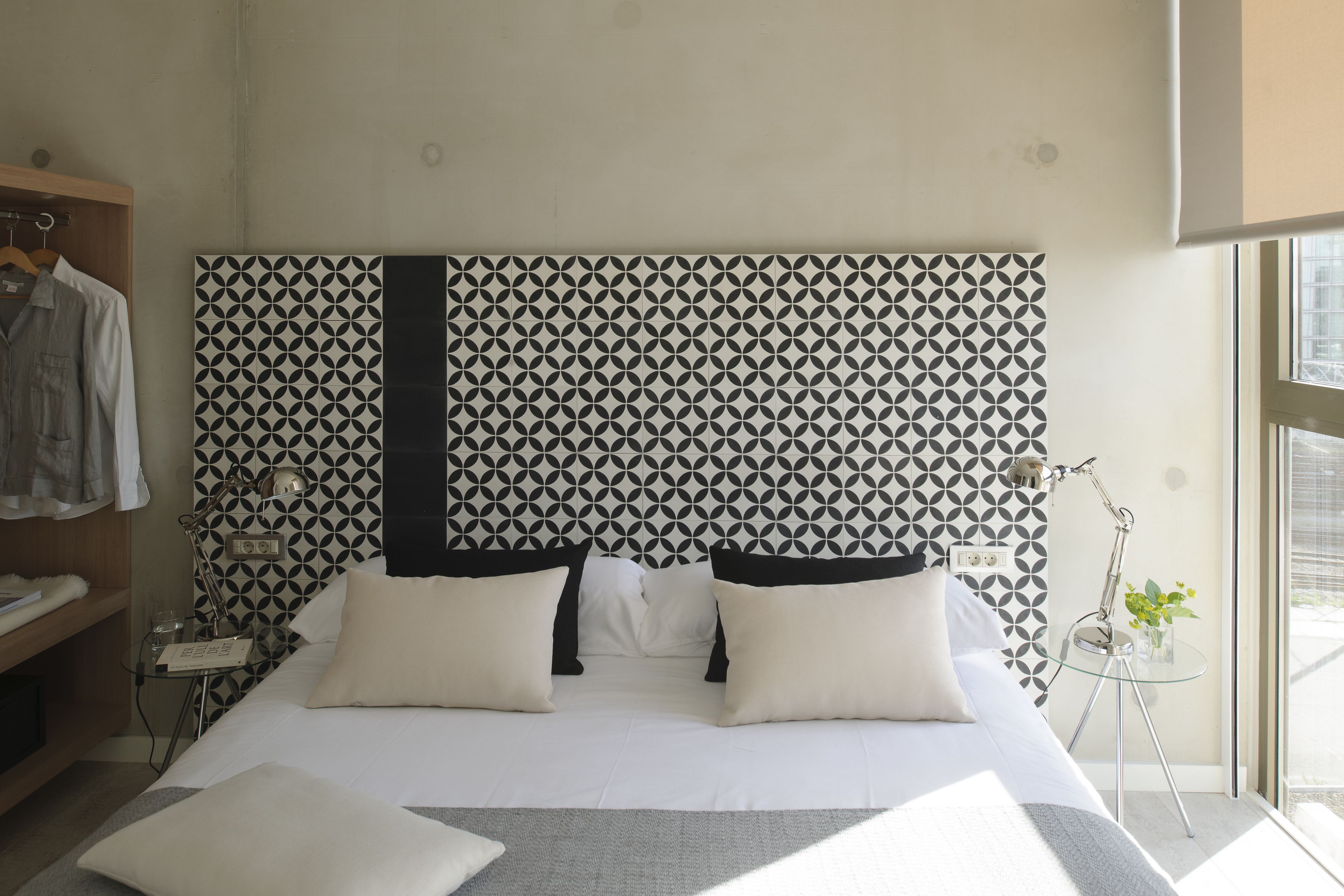 Een moderne hotelkamer met een hoofdeinde met een geometrisch patroon, een net opgemaakt bed met witte lakens, een nachtkastje met een lamp, en uitzicht door een raam.