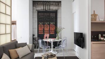 A modern living room with a grey sofa, a small round table, and two clear chairs. A flat-screen TV is mounted on the wall.