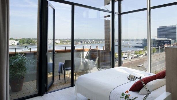 A modern hotel room with large windows, a bed, a desk, and a view of a river and cityscape.