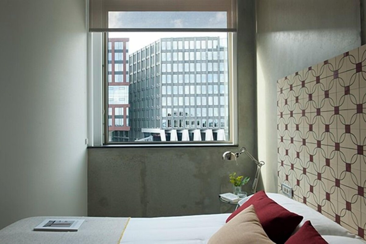 Habitación de hotel con un ventanal grande, una cama con almohadones rojos, una mesita de noche con lámpara y un papel tapiz estampado.