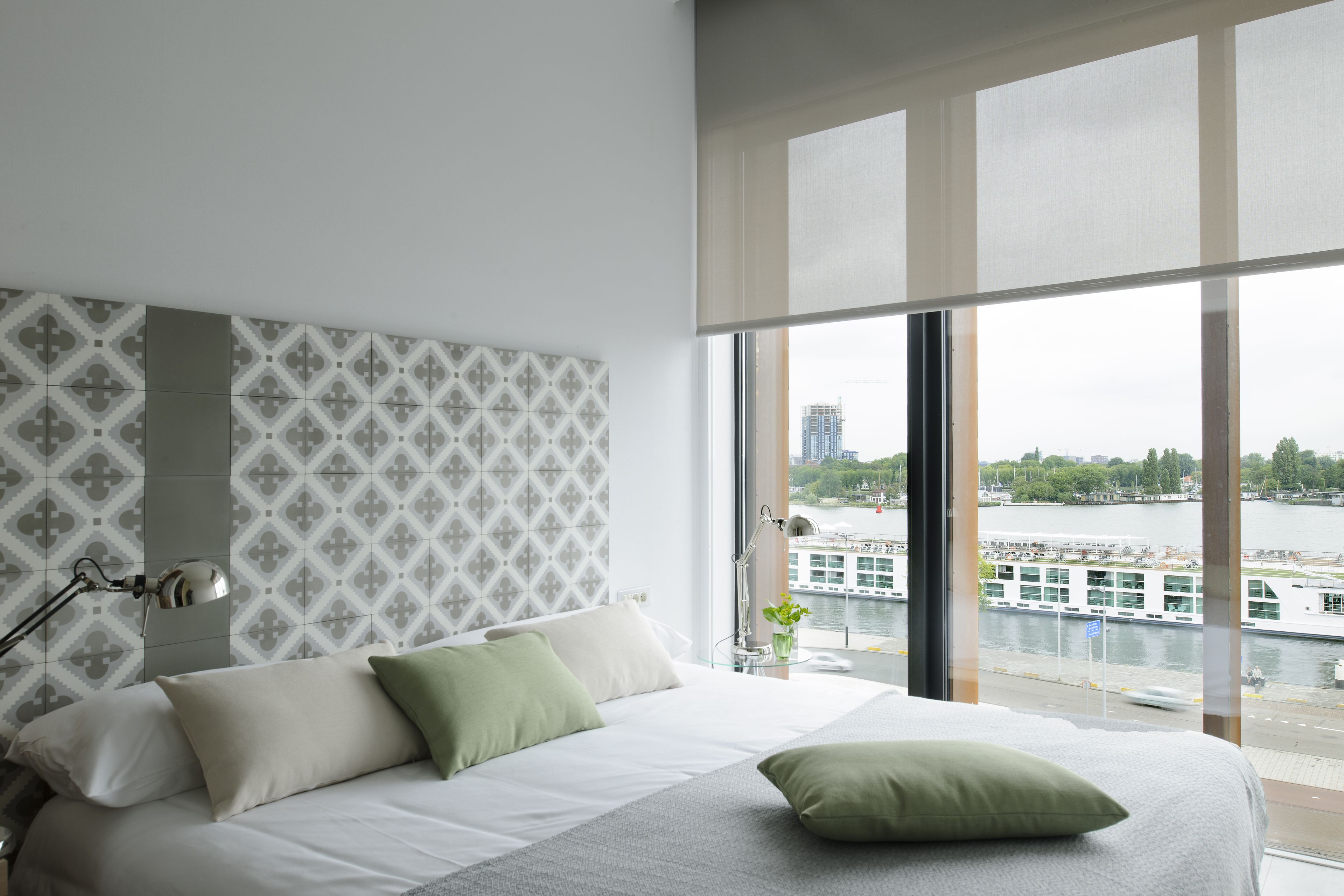 A modern hotel room with a large bed, a nightstand, a lamp, and a view of a river and buildings.
