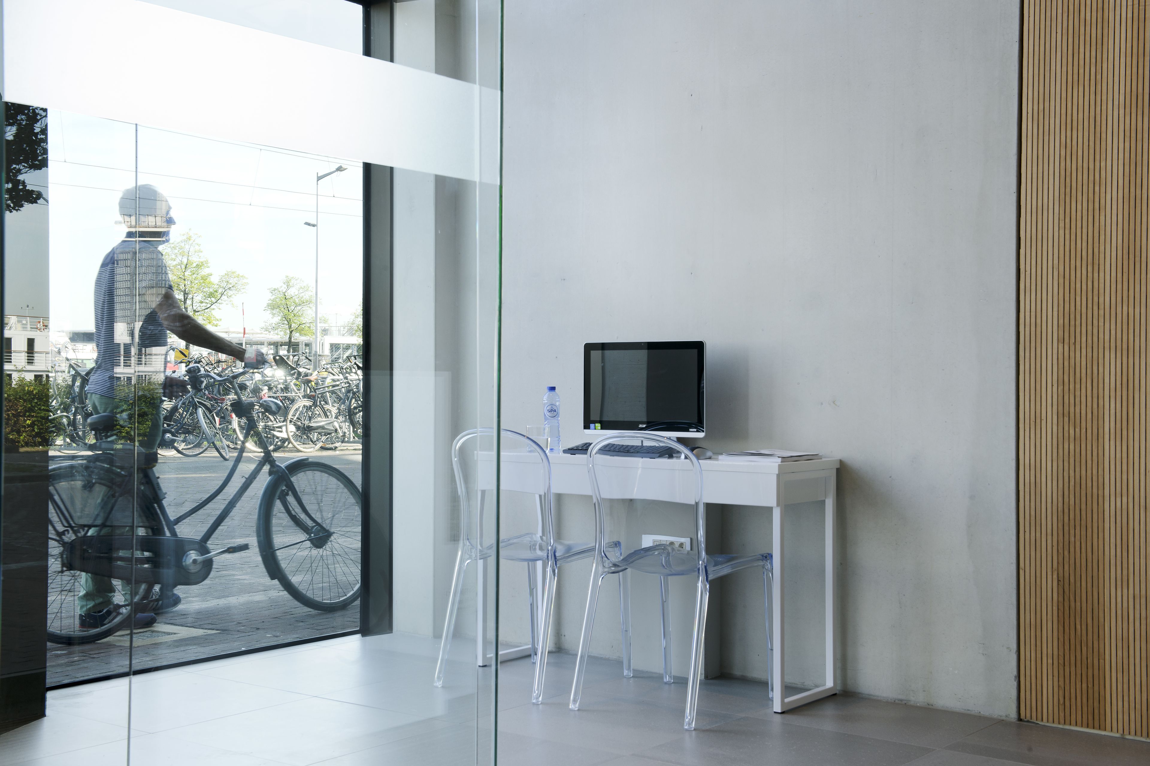 Un espacio de oficina moderno con escritorio, computadora y sillas de plástico transparente. Una bicicleta está estacionada fuera de la ventana.