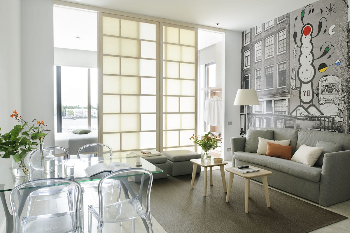 A modern living room with a glass dining table, clear chairs, a grey sofa, a small side table, and a large window with a view of buildings.