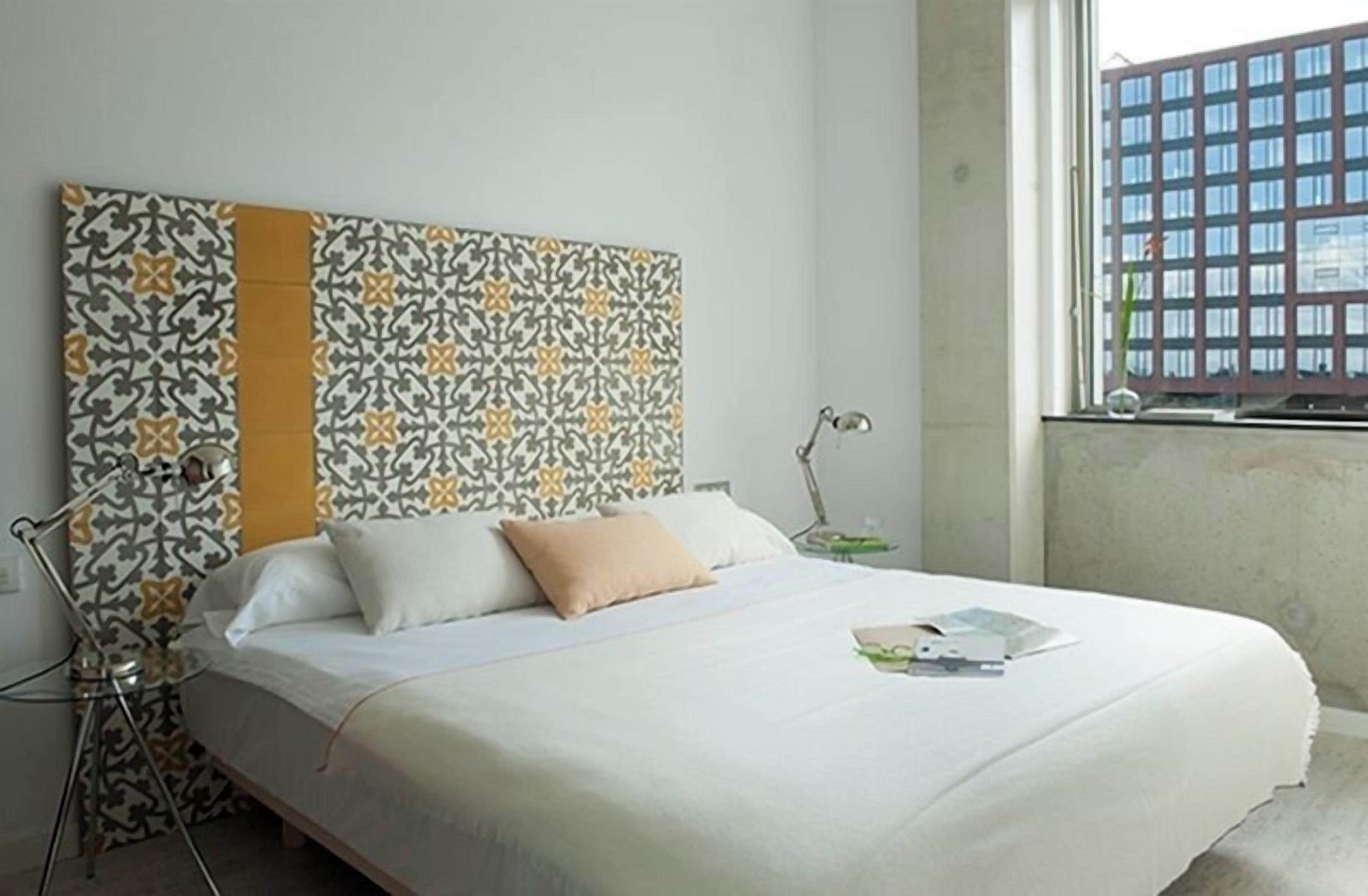 A bed with a patterned headboard, a bedside lamp, and a window with a view of a building.
