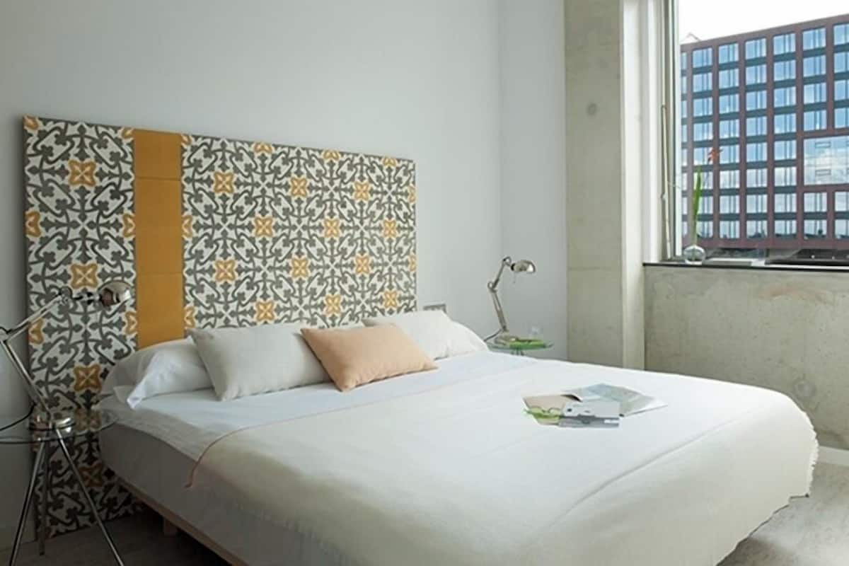 A bed with a patterned headboard, a bedside lamp, and a window with a view of a building.