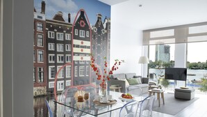 A modern living room with a glass dining table, clear chairs, and a view of a canal and buildings.