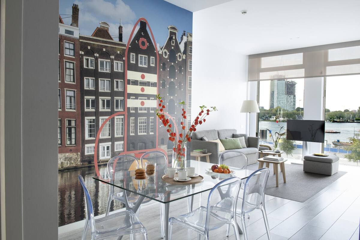A modern living room with a glass dining table, clear chairs, and a view of a canal and buildings.