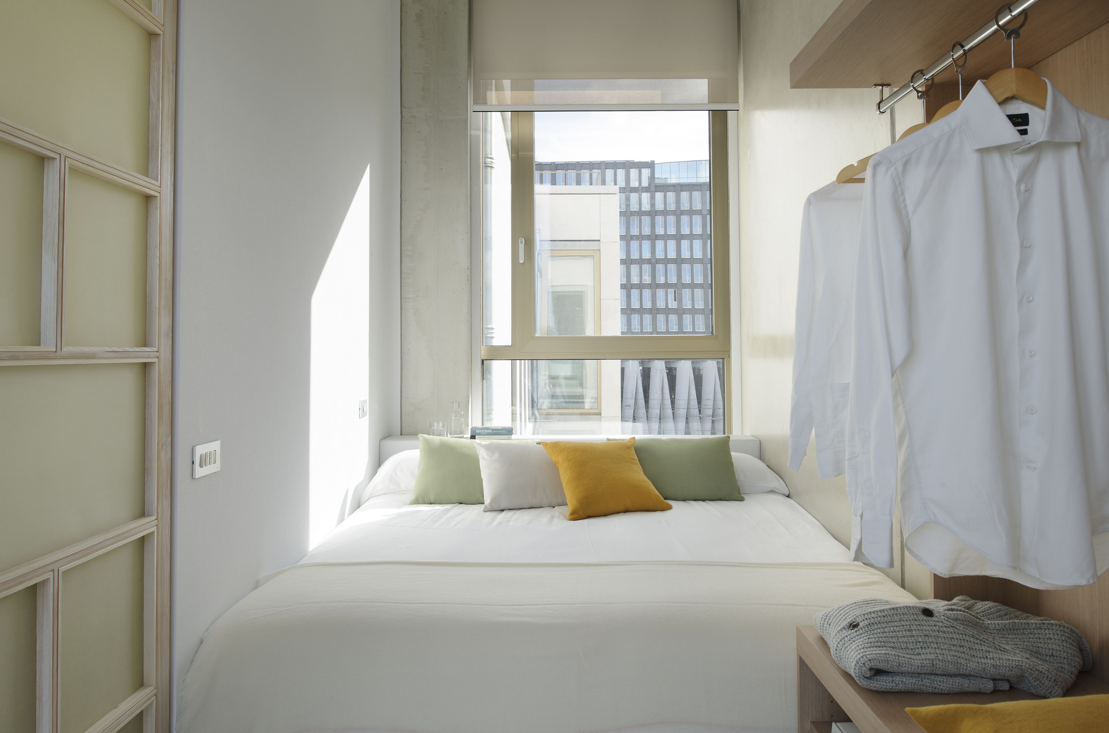 A modern bedroom with a large window, a bed with white bedding, and a wooden bench with a blanket.