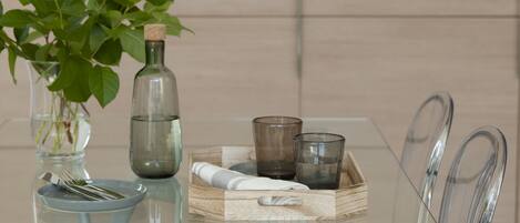 Une table en verre transparent sur laquelle se trouvent un vase rempli de feuilles vertes, une bouteille et deux verres.