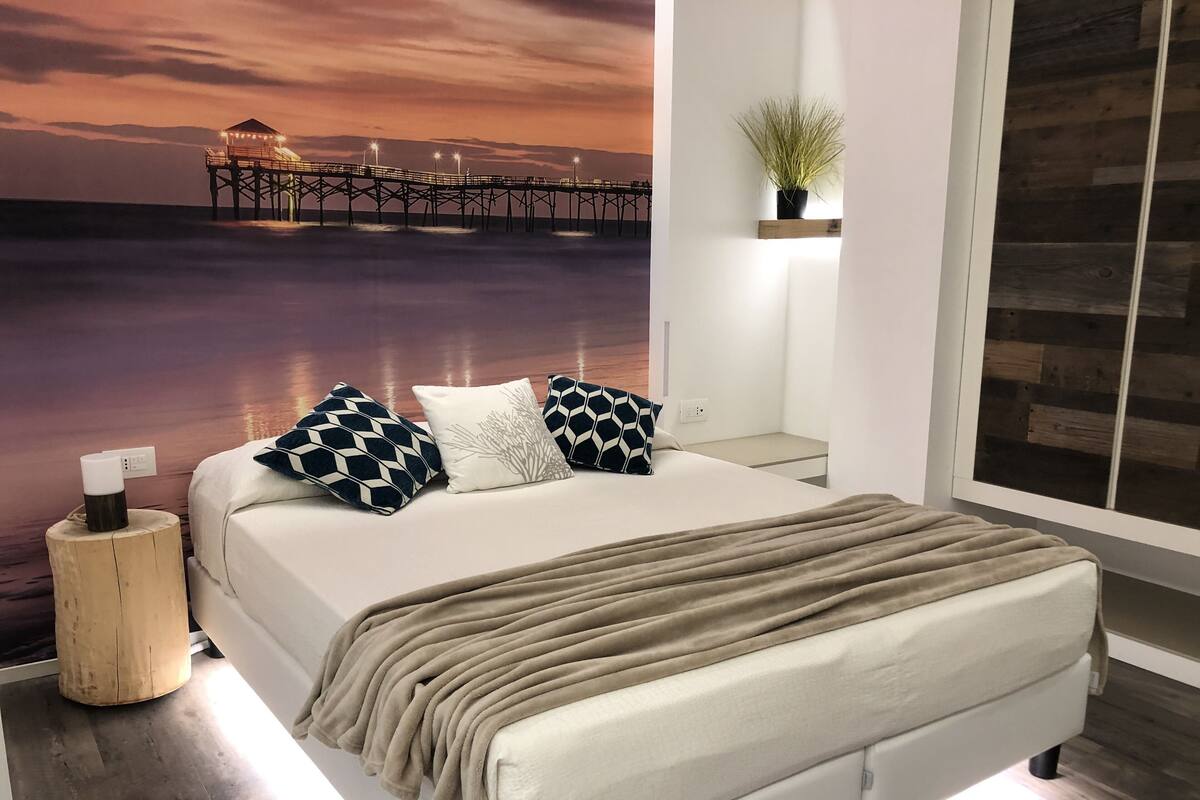 Um quarto moderno com uma cama grande, uma mesinha de cabeceira, uma mesa de apoio com uma luminária e vista para um cais ao pôr do sol.