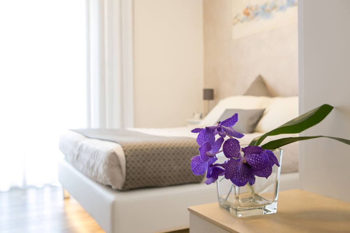 Quarto com cama, mesa de cabeceira e um vaso com flores roxas.