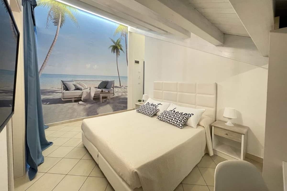 Quarto com cama, mesa de cabeceira, uma cadeira e vista para uma praia com palmeira.