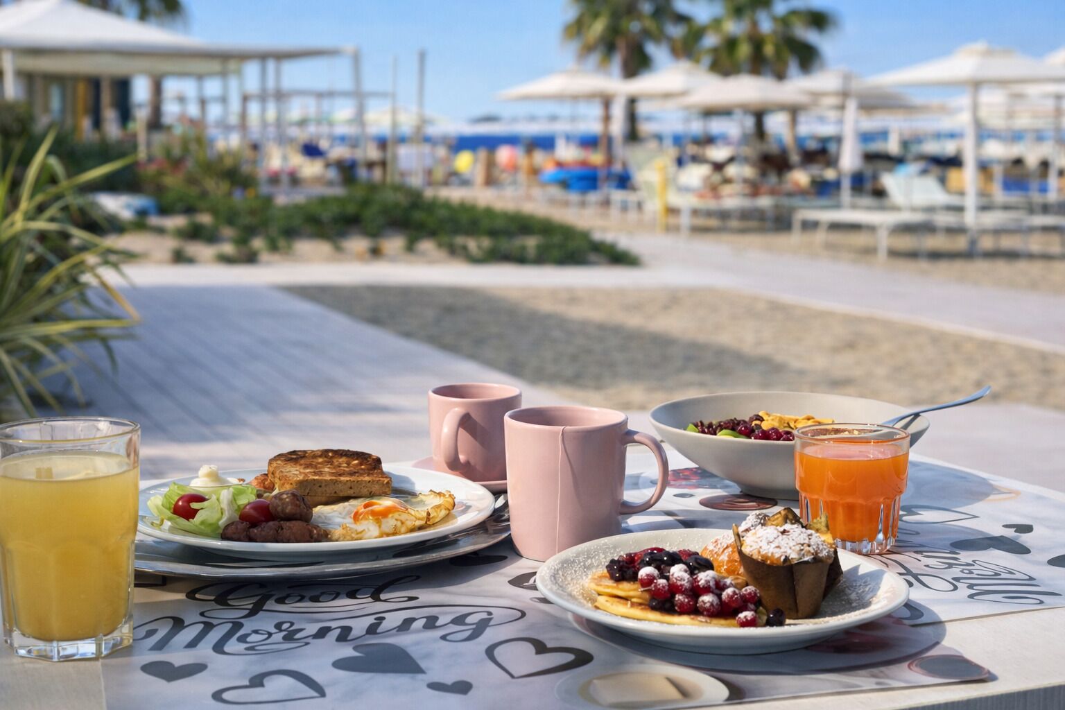 Una mesa de desayuno con tostadas, huevos, bayas y una bebida, frente a una playa con sombrillas de fondo.