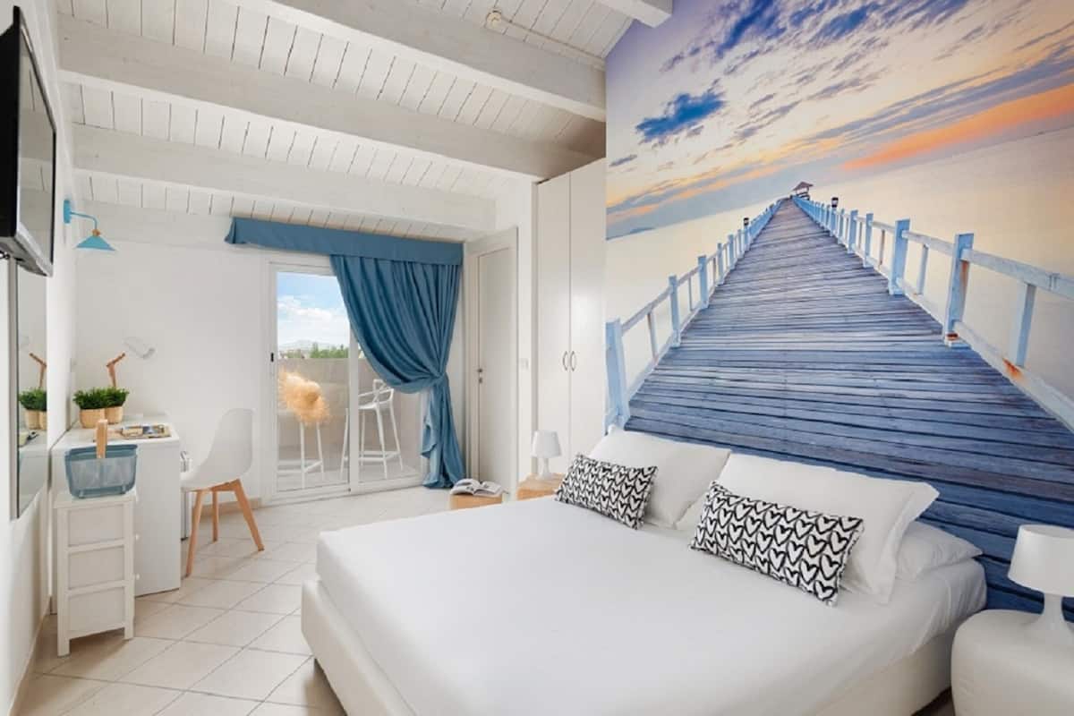 Quarto com uma cama grande, uma escrivaninha e uma cadeira. Vê-se um cais de madeira que leva a um edifício.