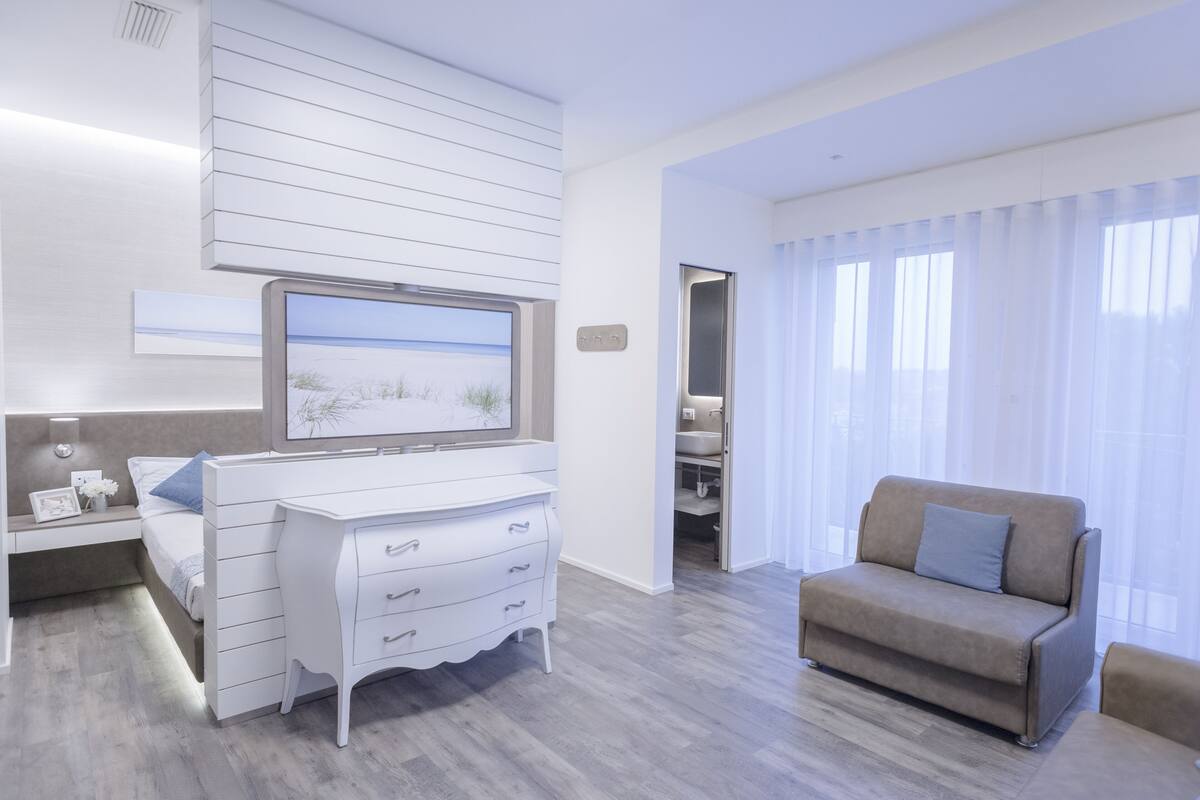 Quarto de hotel moderno com cama, penteadeira branca, televisão na parede e sofá.