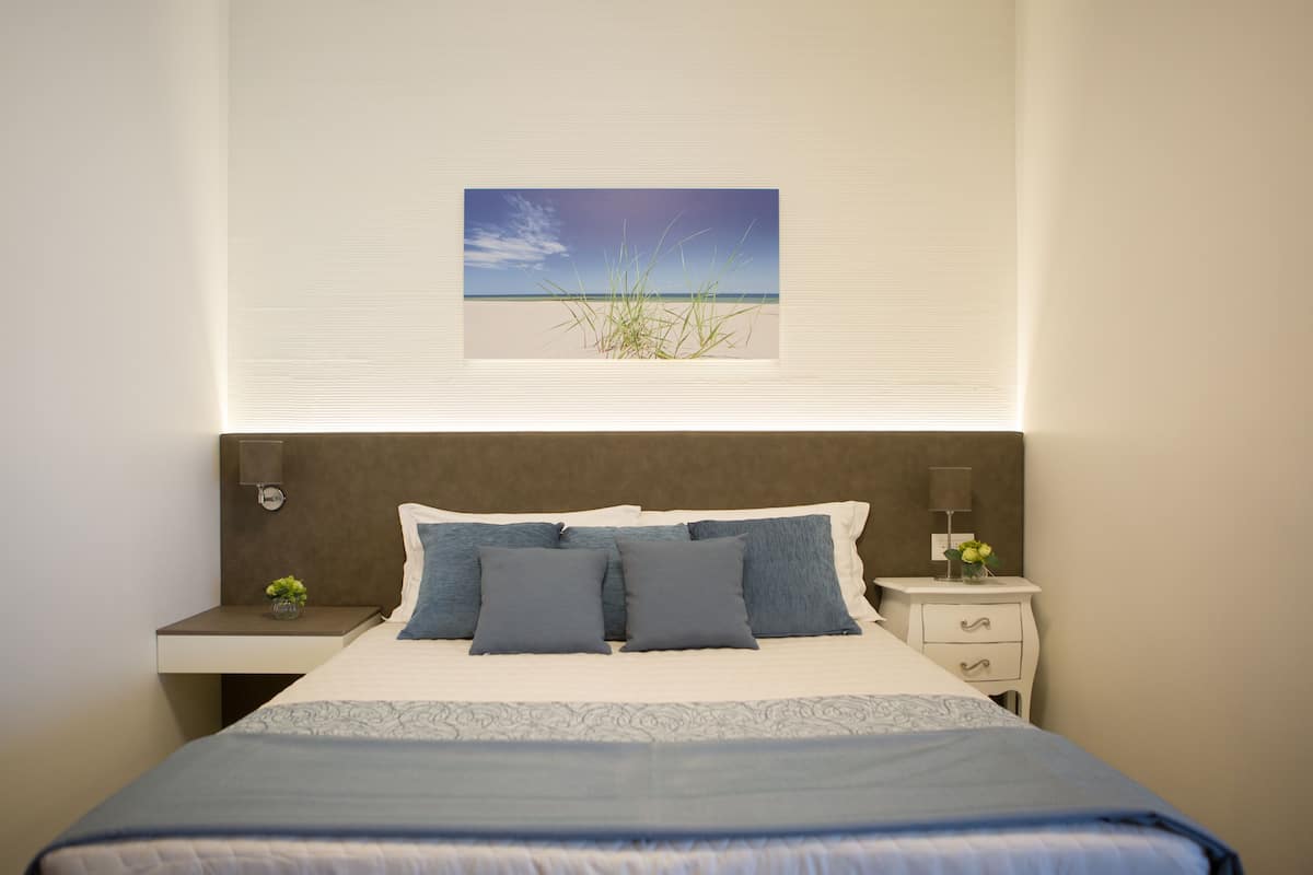 Quarto com cama, mesas de cabeceira, um criado-mudo, uma imagem de praia e uma janela com persianas.