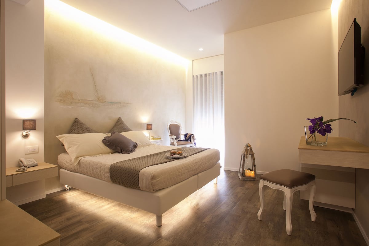 Quarto moderno com cama grande, mesas de cabeceira, um criado-mudo, um pequeno banquinho, abajur e TV fixada na parede.