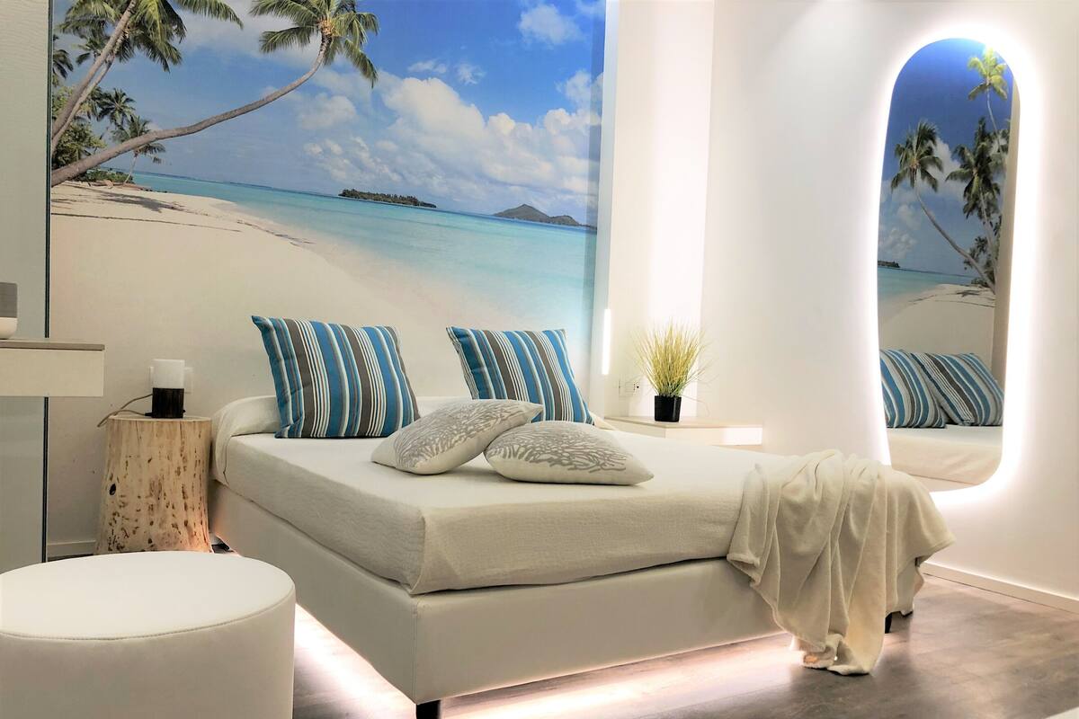 Quarto moderno com uma cama grande, almofadas listradas, uma mesinha de cabeceira com uma planta e vista para uma praia pela janela.