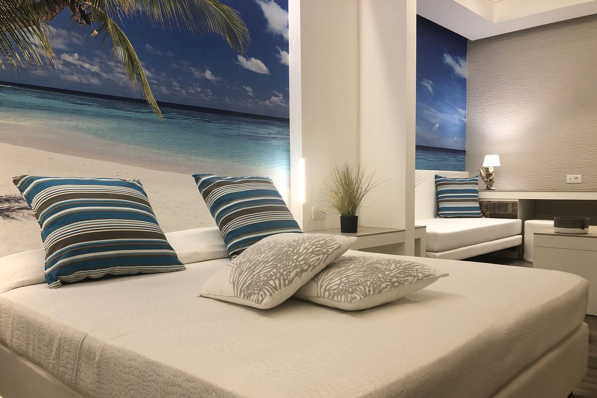 Cama com almofadas listradas, uma planta pequena e vista para a praia e oceano através de uma janela grande.