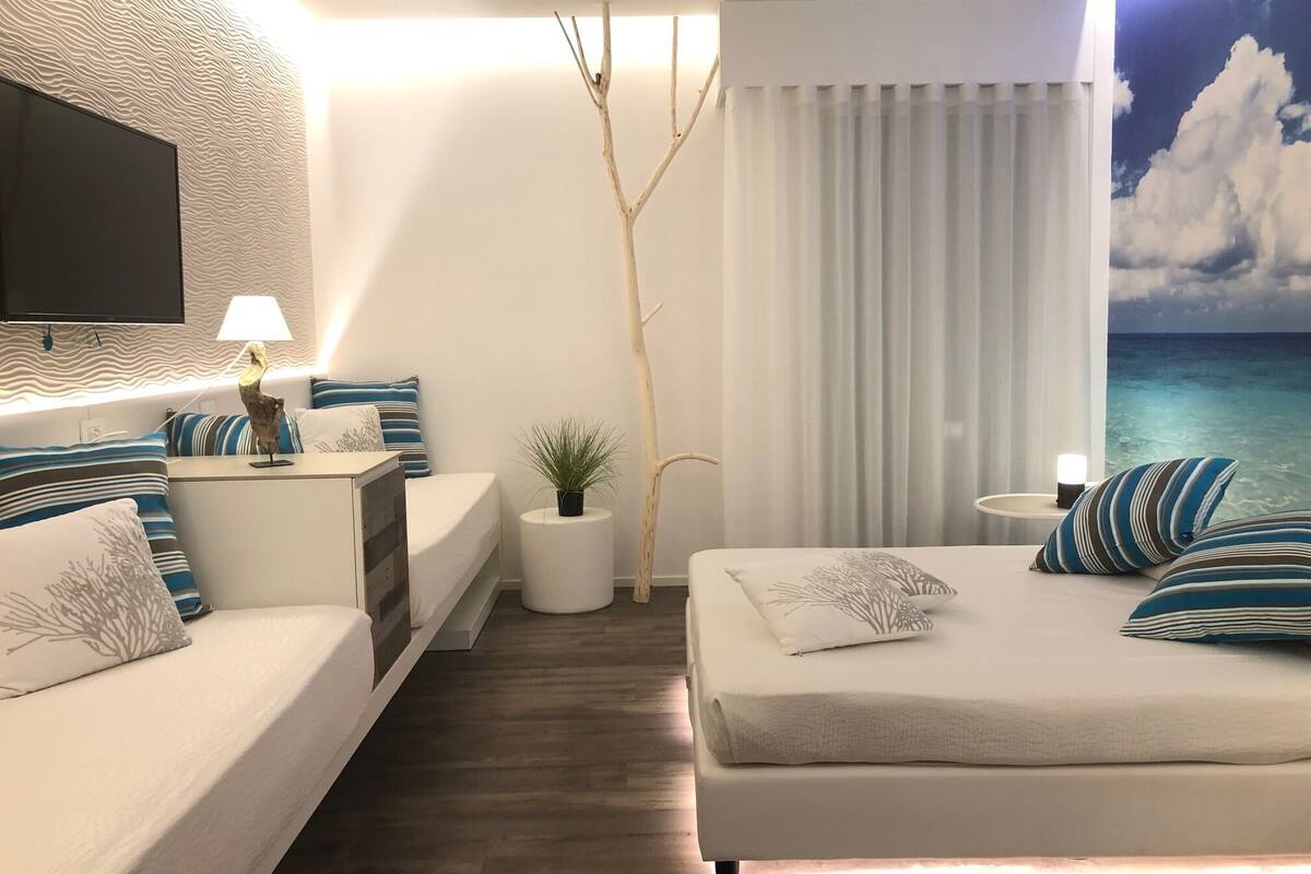 Quarto de hotel moderno com cama, sofá, televisão, luminária, planta e vista para o oceano.