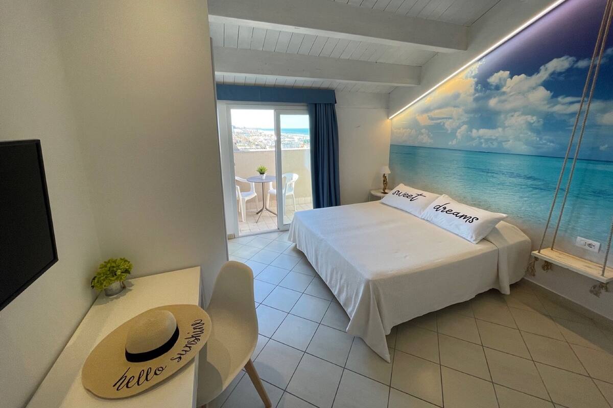 Quarto de hotel com cama, escrivaninha com chapéu, cadeira, mesinha de centro com planta e varanda com vista para o mar.