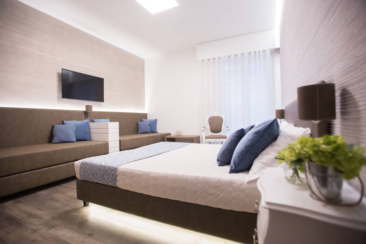 Quarto de hotel moderno com uma cama grande, um sofá, uma TV na parede e uma janela com cortinas.
