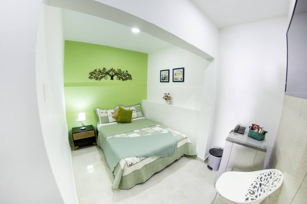A bedroom with a green accent wall, a bed with a quilt, a nightstand, a bedside lamp, a small table with a basket, and a white chair.