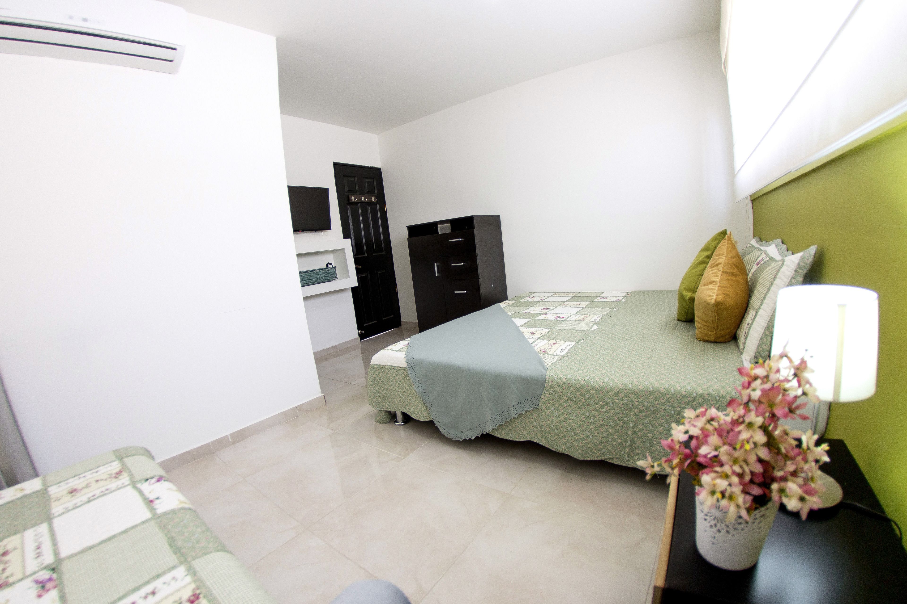 A bedroom with a bed, a nightstand, a lamp, a vase with flowers, a wardrobe, and a door.