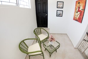 Um corredor com duas cadeiras de vime, uma mesa de centro de vidro com um vaso de flores, uma porta preta e duas quadros na parede.