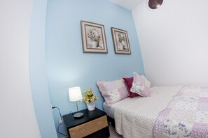 Quarto com parede azul, cama com colcha florida, mesa de cabeceira com abajur e um vaso de flores, e duas fotos emolduradas na parede.