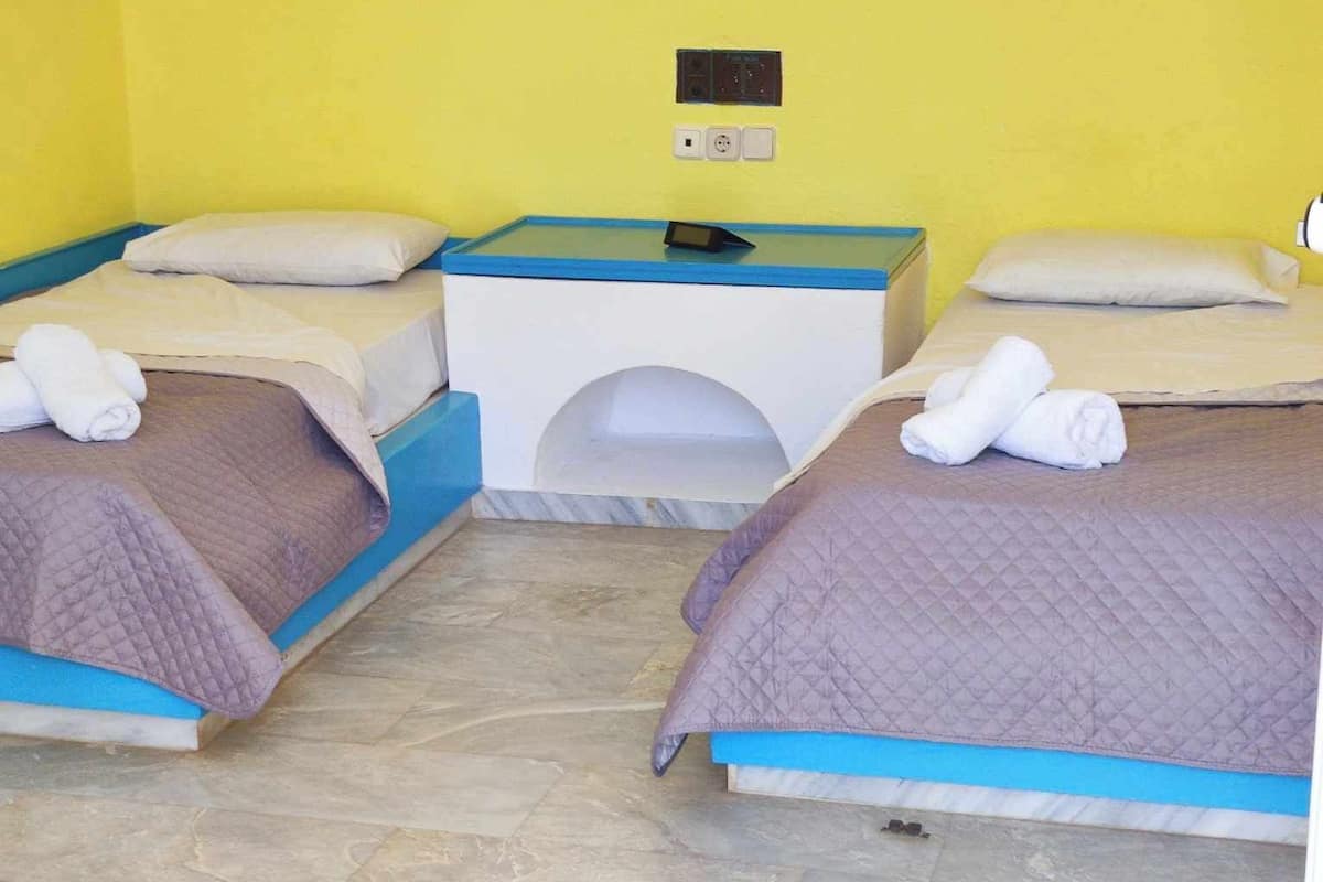 A hotel room with two beds, each with a blue headboard, a small table with a lamp, and a wall-mounted control panel.