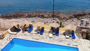 A pool area with blue lounge chairs and umbrellas, a clear view of the sea, and a rocky shoreline. - Sissi Mare Apartments (Agios Nikolaos)
