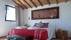 A bedroom with a wooden ceiling, a bed with red bedding, a colorful rug, a wooden nightstand, a lamp, and a vintage trunk. - Herdade da Maxuqueira (Gaviao)