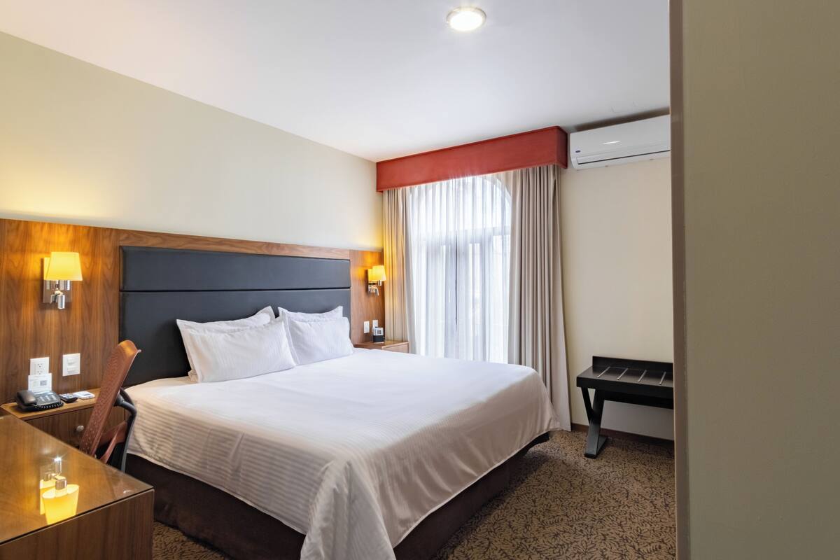 A hotel room with a large bed, bedside tables, a desk, and a window with curtains.