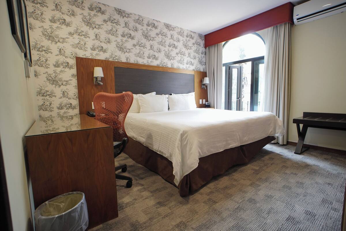 A hotel room with a large bed, a wooden headboard, a desk with a chair, a trash bin, and a window with curtains.
